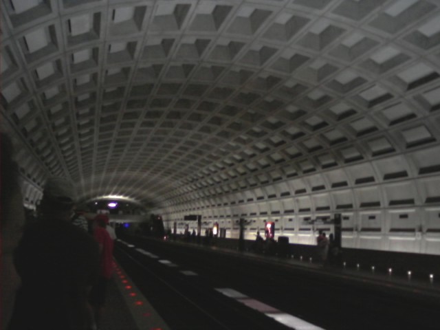 A photograph taken from the platform of a Metro station in Washington, D.C. looking down the track. The iconic waffle-like arched design of the Metro system is clearly visible, with the straight lines converging toward a vanishing point.